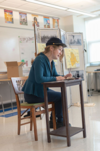 一个学生在教室里, sitting in a chair at a small desk with a pen in h和, 身体前倾, 表演 